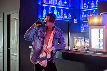 Young man drinking a beer inside a bar with neon lights in the background.