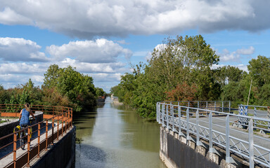 Pedestrian bridge over a narrow canal in Marans, France, captured in late summer. The wooden walkway with metal railings crosses calm water, surrounded by green trees and a clear sky in a quaint river