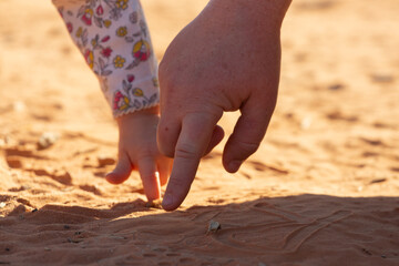 The hands of a woman and small child draw in the red sand. The woman is showing the child how to make letters.