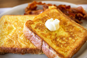 Two pieces of French Toast on a white plate with bacon blurred in the background.  Daub of butter melts ontop.  Diner shot.