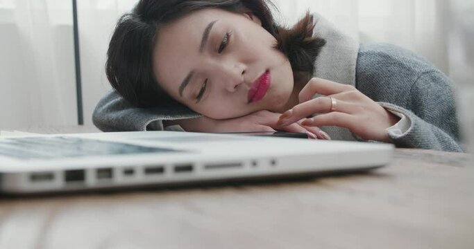 very borin young asian woman in a gray coat leaning over the table after working on laptop playing mobile phone business freelance woman tired