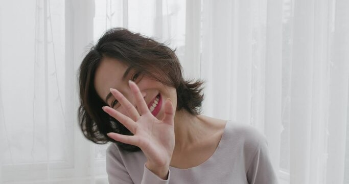 Young Asian woman in a gray T-shirt standing in front of a white curtain talking to camera pretty cute lovely lady chatting speak to webcam indoor POV view of happy girlfriend talk 