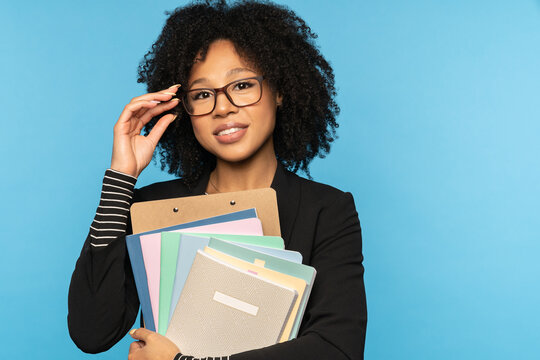 Happy teacher or businesswoman in blazer, wear glasses, holding notebooks, documents and folder,  smiling looking at camera isolated on blue studio wall. Education in high school university college 