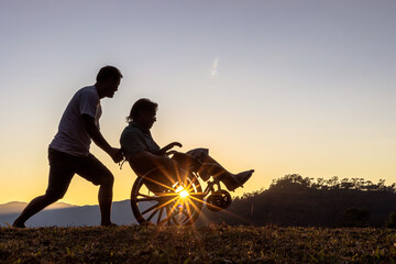 Silhouette of joyful disabled man in wheelchair running playing with friend at sunset