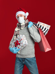 A young man in a medical mask and gloves, wearing a Santa hat and scarf holds a gift box and shopping bags on a red background