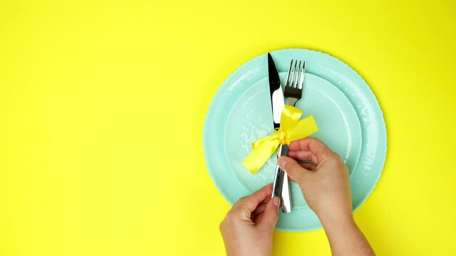 two female hands put metal knife and fork tied with yellow ribbon on empty plates, top view, yellow background, table setting