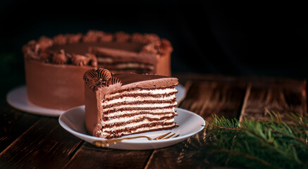 Chocolate cake on a white plate. Against the background of a branch of a Christmas tree. Wooden table with a beautiful pattern. Traditional sweets for birthday and other holidays
