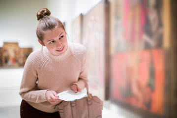 Portrait of young pensive female visitor with guide book looking at exhibition in art museum