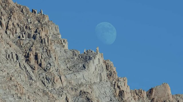 Large moon sits right behind rugged rocky mountain with a deep blue sky during the day (60 fps slowed to 30 fps)