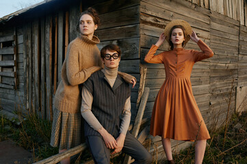 Portrait of three young beautiful people in village area. Wooden farm. Backyard. Nature. Yellow grass. Sunset light, autumn. Country style clothes