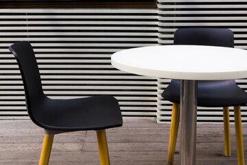 A white table and two black chairs putting next to a wooden wall outside a cafe. 