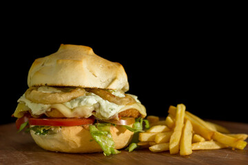 Close up of a homemade burger with onion rings on a chopping wood board, on black background