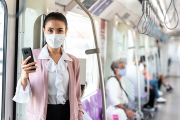 Asian business woman travelling on train in city wearing mask and pink blazer use smartphone