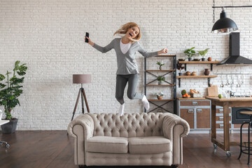 Cheerful positive woman jumping on couch in the air, raised hands with happiness