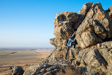 A girl with a backpack climbs a high rock above the plain in summer at the Golden hour.