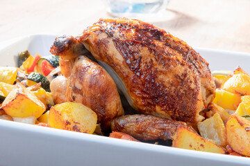 close up of a spit-roasted chicken with baked potatoes and assorted vegetables on a baking dish