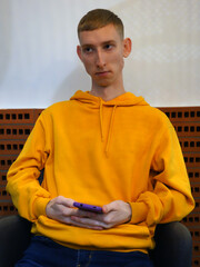 Handsome young guy holding a phone and looking around. A guy in a casual yellow hoodie sitting on a chair.
