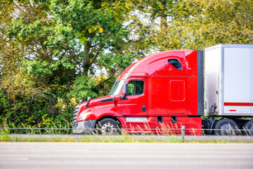 Fototapeta na wymiar Bright red big rig semi truck with dry van semi trailer running quickly on the highway with green trees on the side for timely delivery