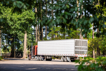 Brown classic big rig semi truck with reefer semi trailer driving on the exit form rest area parking lot road in green forest