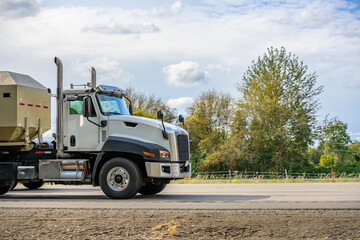 Day cab white big rig semi truck with industrial dump semi trailer running on the highway road to working place destination