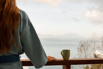 Cropped image woman in bath robe looking at beautiful ocean view from a tropical balcony with morning coffee. Focus ob the cup