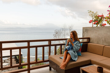 Young woman wearing blue bath robe sit on the coach hotel's balcony looks at the sea holding cup of coffee in hands