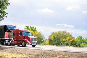 Fototapeta na wymiar Red big rig semi truck transporting covered semi truck with front wall spoiler running on the road