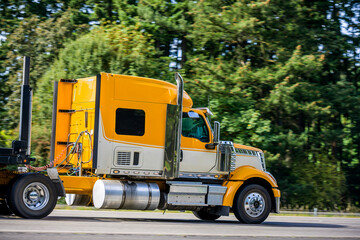 Side of big rig yellow semi truck with semi trailer running on the flat road with green trees on the shoulder