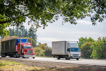 Two different carriers red big rig semi truck with dry van semi trailer and small rig white cab over truck with box trailer running on the highway side by side