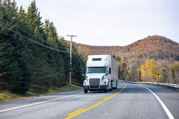White big rig semi truck transporting cargo in dry van semi trailer running on the winding autumn road in New England