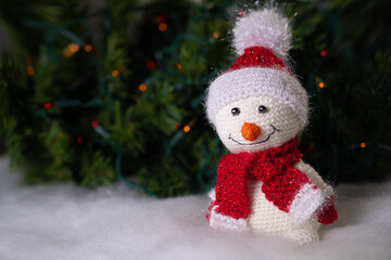 A small cute toy snowman made with his own hands in a Santa Claus hat stands on the snow on the background of a Christmas tree: place for text, Christmas background