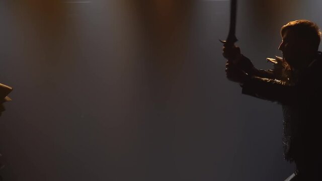 Men fight with swords in a dark studio with stage lighting. Silhouettes of male actors in armor performing in a theatrical show. Close up in slow motion.