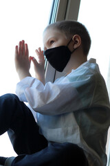 A little boy in a black mask at the window, self-isolation and quarantine, coronavirus.