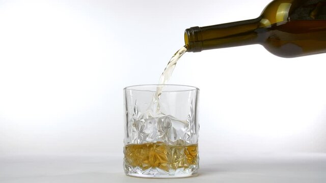 Golden whiskey poured into a crystal clear glass from a brown bottle. Whiskey/hard drink pouring from a bottle into a transparent glass with ice cubes against a white background