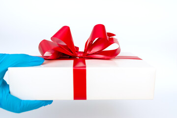 Holiday gift.Hand wearing meidical gloves holding gift box with red ribbon.white background.