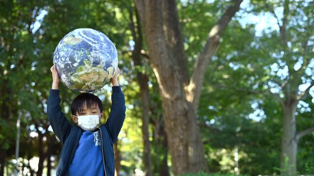 マスクをした男の子が地球の形をしたボールを持ち上げている。covid-19イメージ。