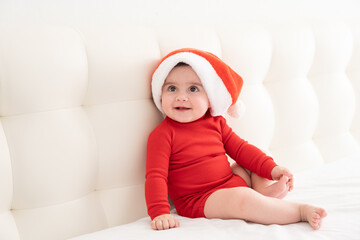 baby girl 6 month in red bodysuit and Santa hat sitting on bed. minimalist.