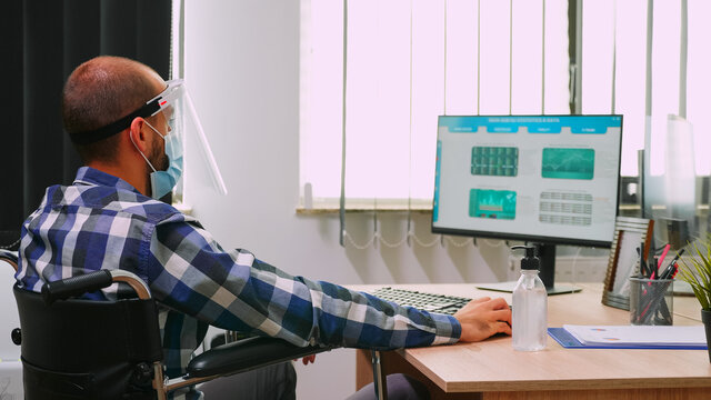 Disabled businessman sitting in wheelchair wearing face mask and visor against coronavirus disinfecting hands before checking financial data in business office. Freelancer respecting social distance