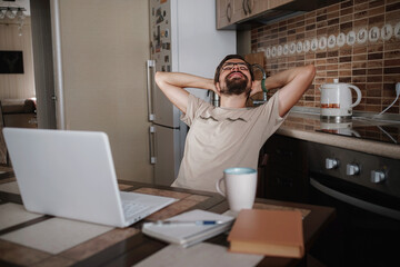 Smiling hipster freelancer holding hands behind head sitting at office desk behind laptop.