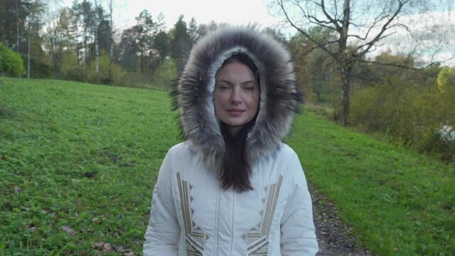 Beautiful woman with brown hair walking in park on cold autumn day with trees covered in yellow leaves in warm fur collar coat