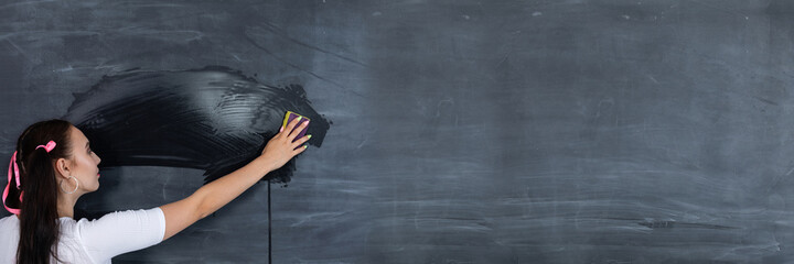 In a panoramic shot of a standing teenager wiping school blackboard with a wet sponge. The school blackboard is hanging on a white brick wall. A helpful girl stands at the blackboard.