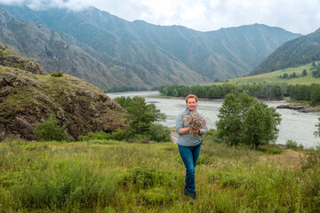 Woman in a singlet and jeans with a bouquet of wild flowers on the river bank against the mountains and cloudy sky