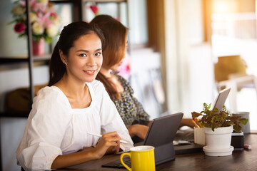 Two beautiful Asian women Sitting at work online or studying during the corona virus with a tablet in the morning, the sun warms happily.