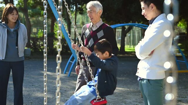 公園でブランコに乗る子供を見守る両親と、ブランコを押すおじいさん