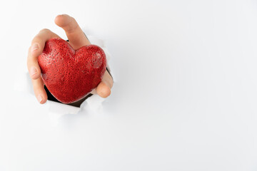 A woman's hand holds a heart and breaks through a white wall. A red heart pierces the paper.