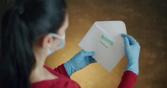 Woman Opening Envelope with Certificate with Positive Results in Covid-19. Over Shoulder View of Woman Wearing Surgical Gloves Opening Envelope and Pulling Out Positive Test Results in Coronavirus.