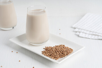 Gluten-free and lactose-free healthy buckwheat milk full of vitamins, minerals and antioxidants served in drinking glass on plate with grains and textile towel on white wooden background. Horizontal