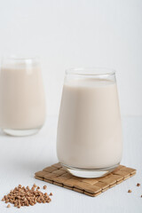 Gluten-free and lactose-free healthy buckwheat milk full of vitamins, minerals and antioxidants served in tall drinking glass on wooden mat with raw grain ingredient on white background. Vertical