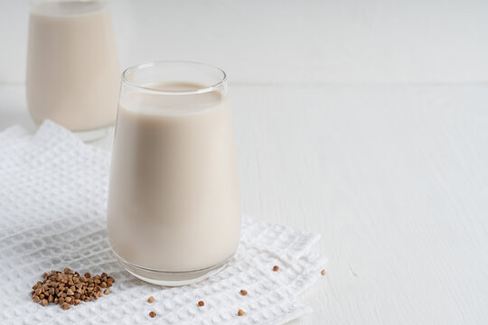 Gluten-free and lactose-free healthy buckwheat milk full of vitamins, minerals and antioxidants served in drinking glass on textile towel on white wooden background at kitchen. Image with copy space