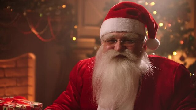old kind Santa is sitting in his room in residence at Christmas holiday, holding finger near lips to show keep silence and secret